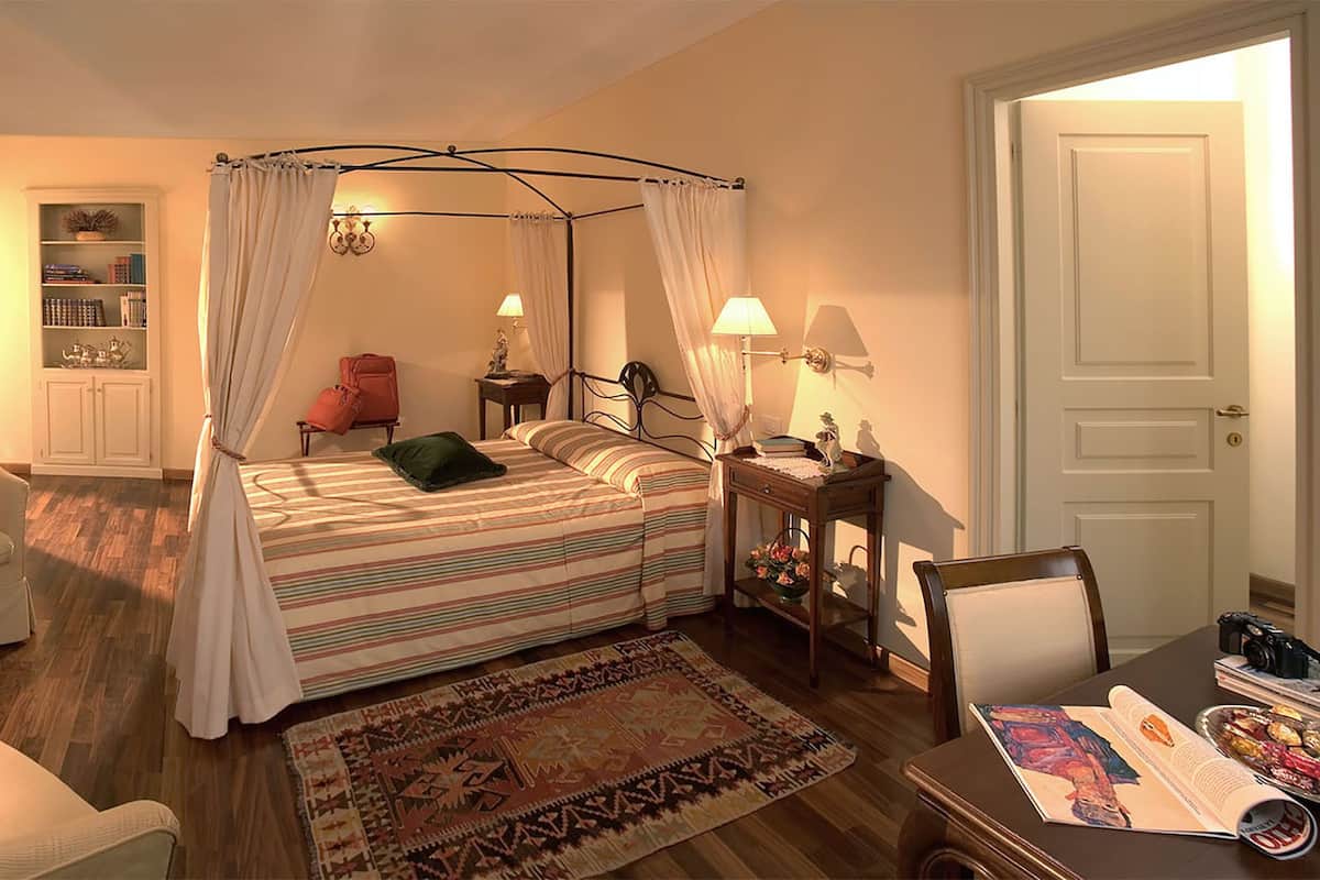 A cozy bedroom with a four-poster bed, a fireplace, a bookshelf, and a bedside table with a lamp.