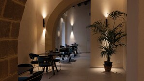 A corridor with tables and chairs, a potted plant, and wall-mounted lighting.