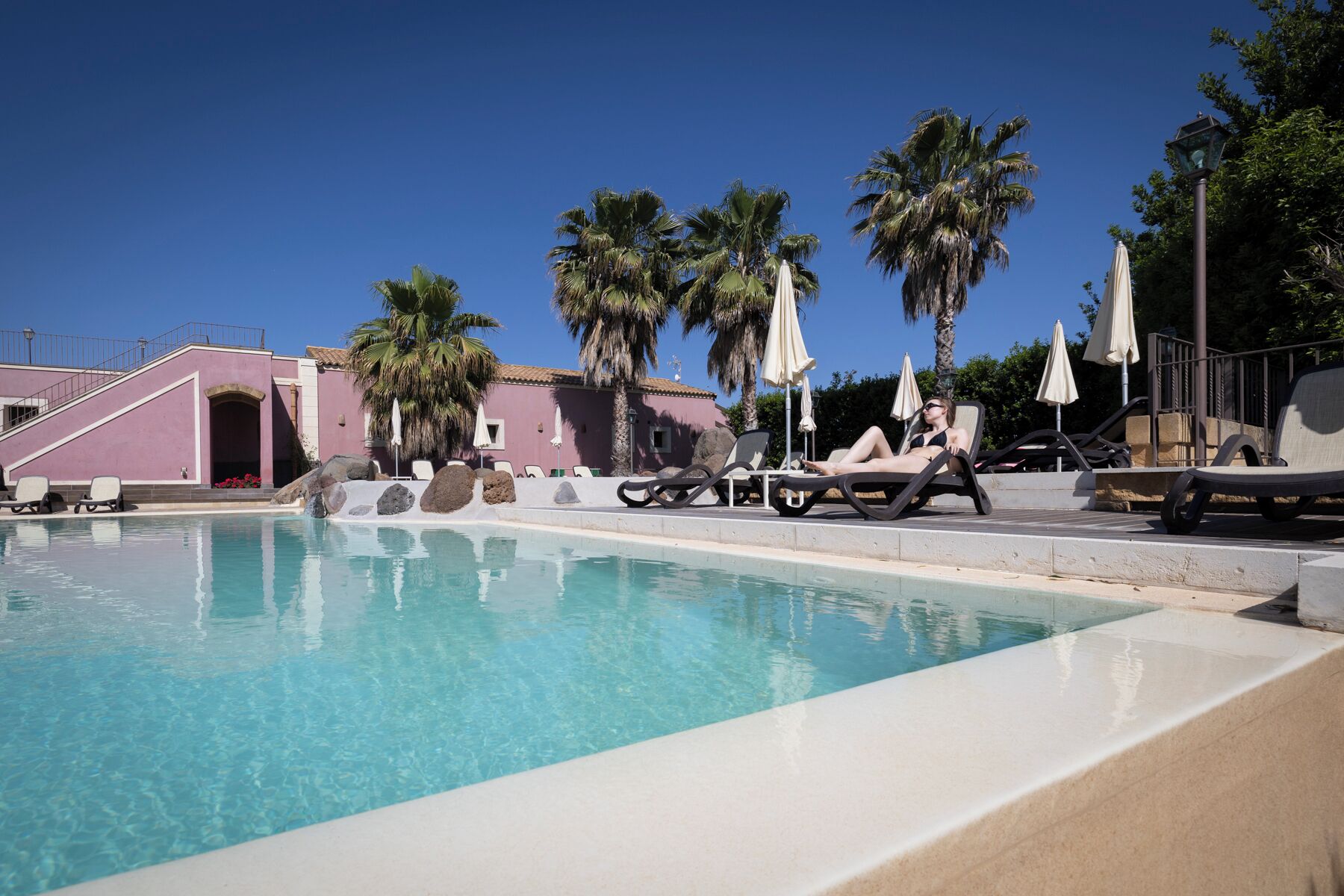 A pool area with lounge chairs and umbrellas, a pink building, and palm trees.