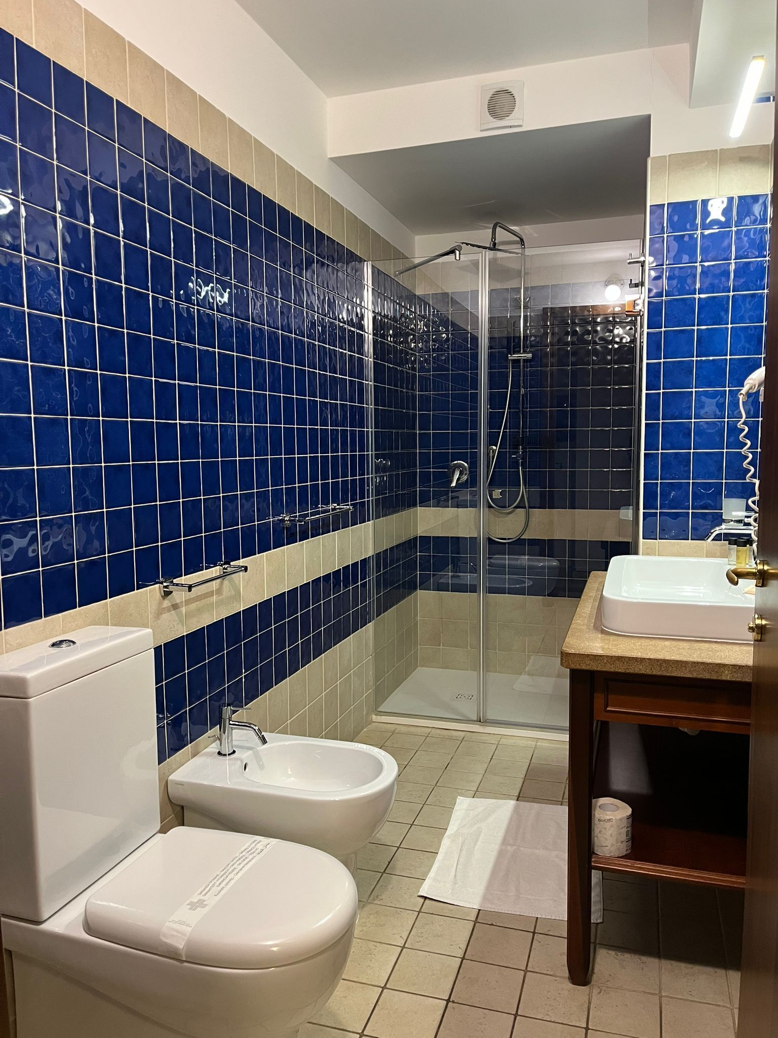 A bathroom with blue-tiled walls, a glass-enclosed shower, a white sink, and two toilets.