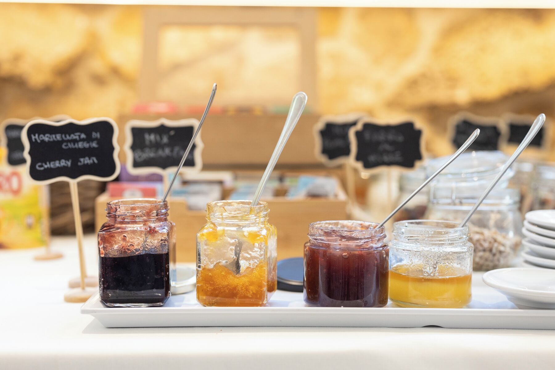Variedad de frascos con etiquetas que señalan distintos tipos de mermeladas y salsas, junto a cucharas metálicas.