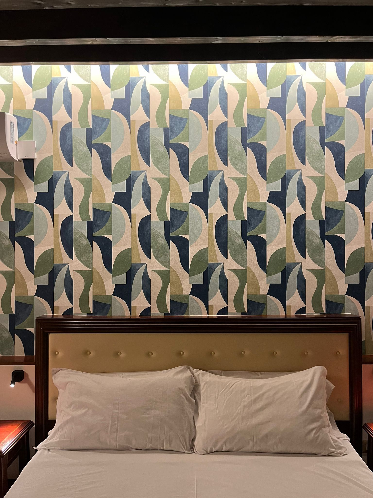 Un lit doté d’une tête de lit capitonnée, un mur orné d’un motif géométrique et une table de chevet avec une lampe.