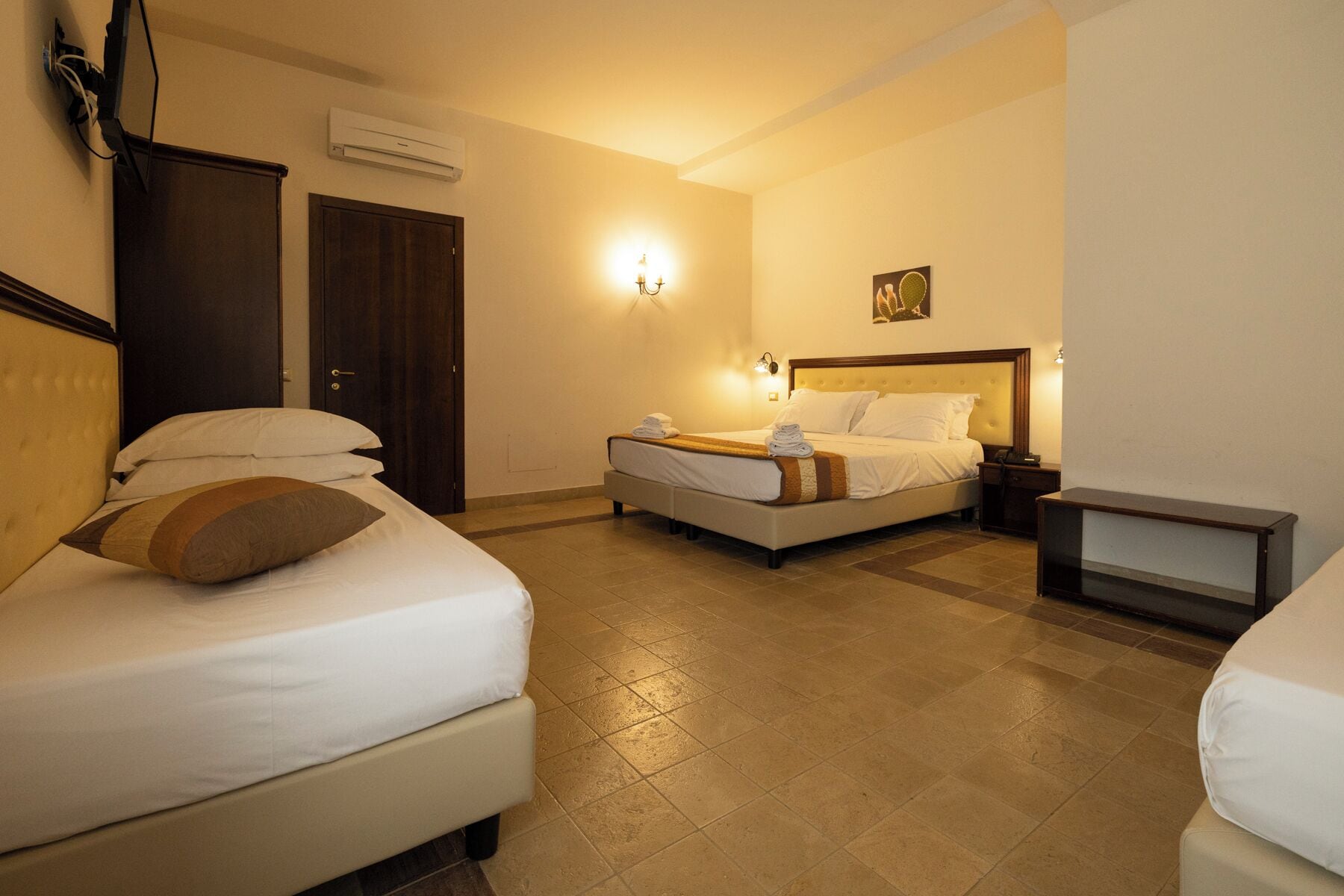 A hotel room with two beds, a bedside table, a nightstand, an air conditioning unit, and a wall-mounted light fixture.