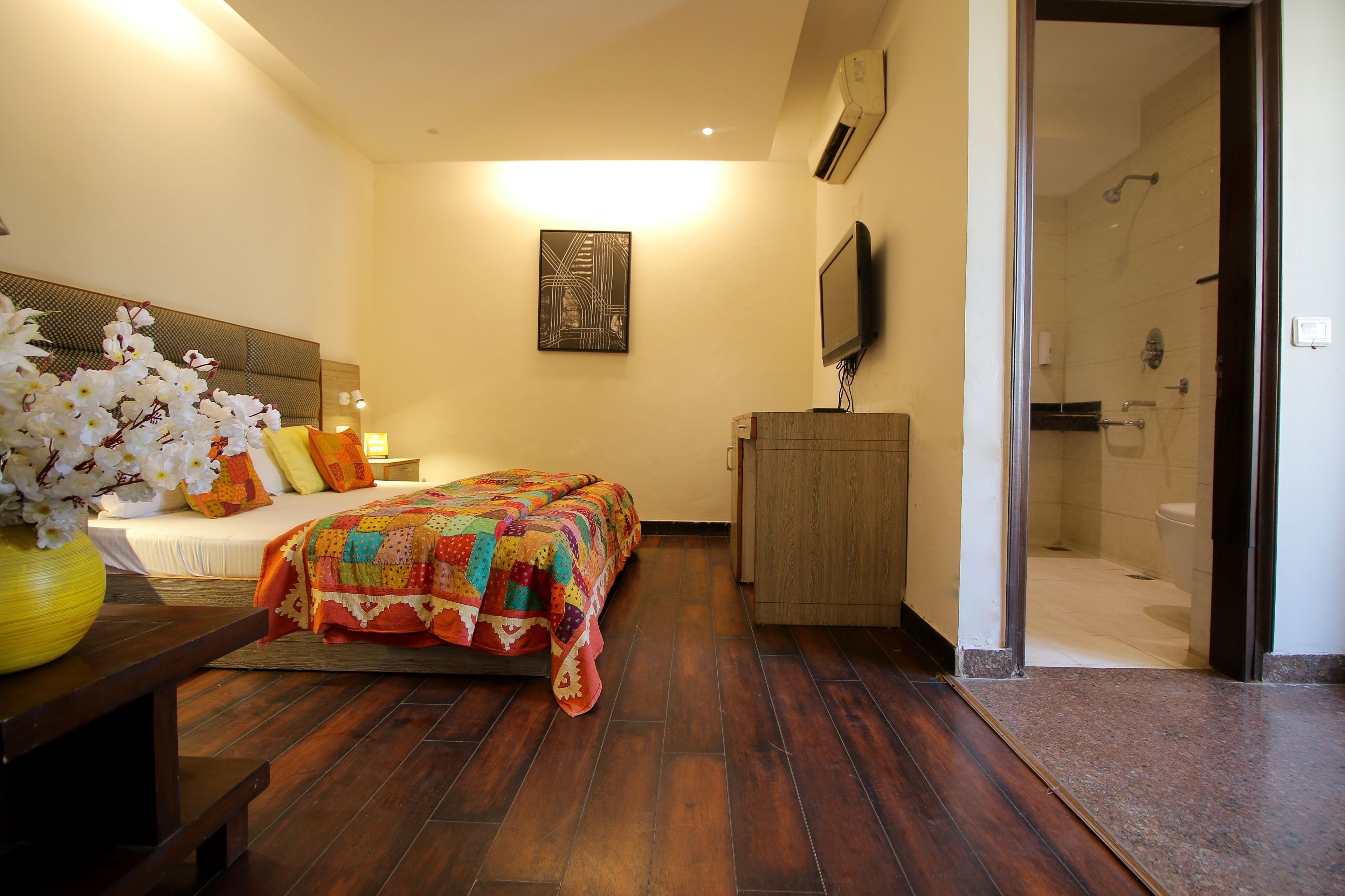 A hotel room with a bed, a wooden nightstand, a TV, a shower area, and a wall-mounted air conditioner.