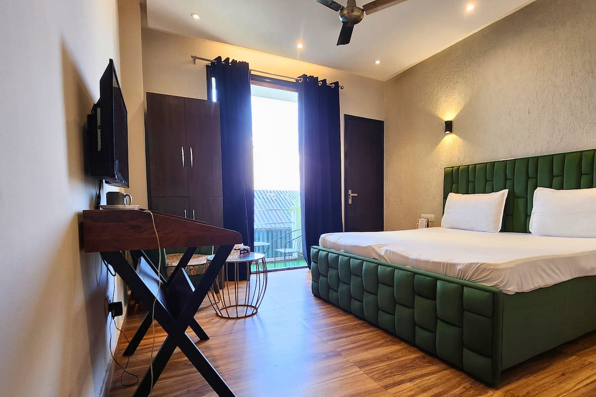 A modern hotel room with a green upholstered bed, wooden flooring, a desk with a television, and a view of the outdoors through a large window.