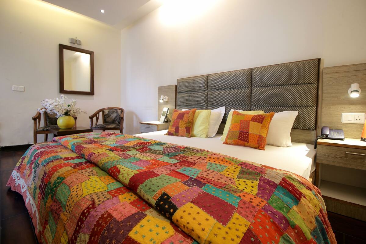 A neatly made bed with a colorful quilt, a bedside table with a lamp, and a chair with a vase of flowers.