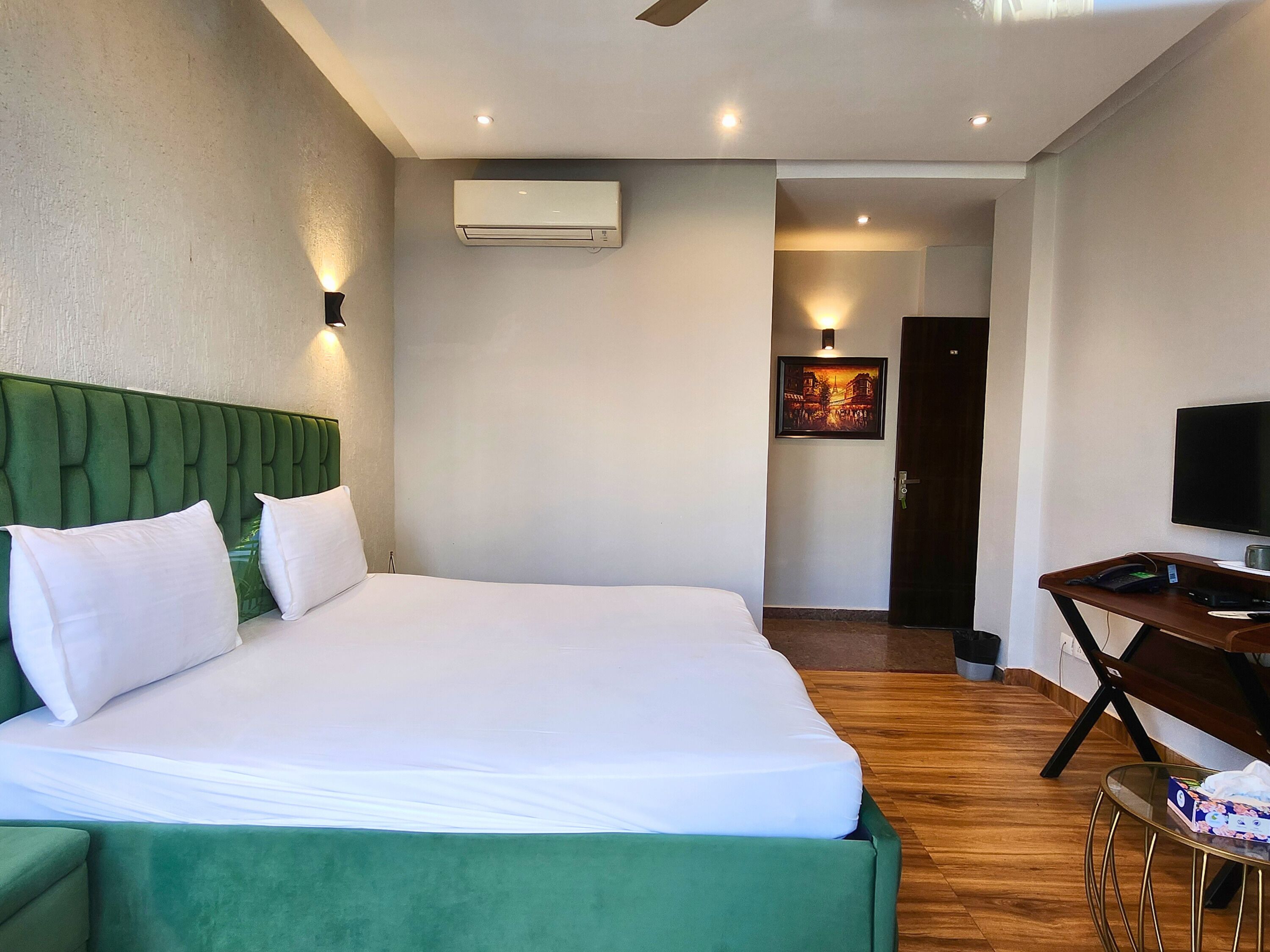 A hotel room with a large bed, a desk with a chair, a flat-screen TV, and an air conditioning unit on the wall.