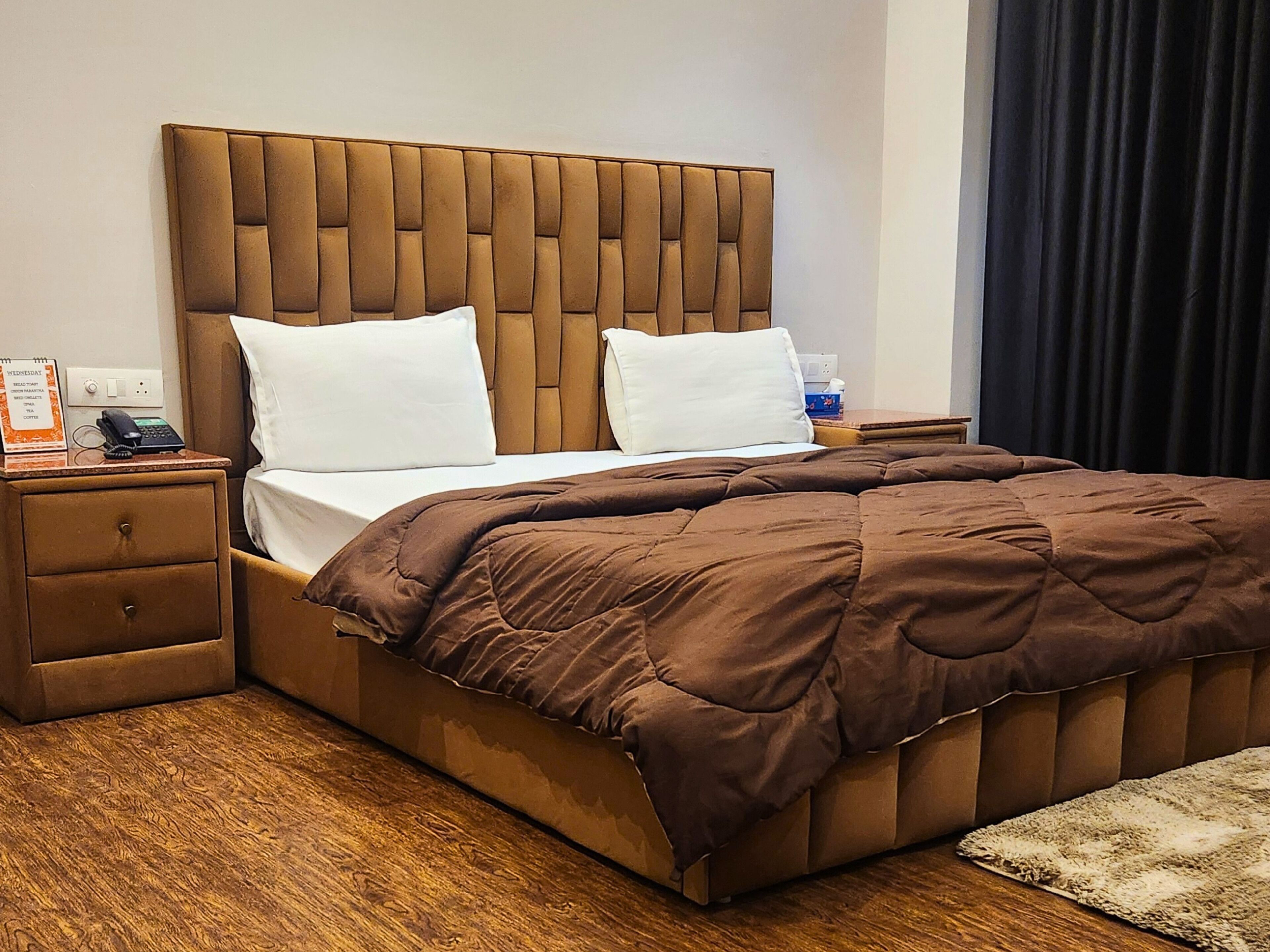 Habitación de hotel con cama tapizada en tonos marrones, almohadas blancas, colcha marrón, mesita de noche de madera con teléfono y una pequeña mesa con una caja de pañuelos.