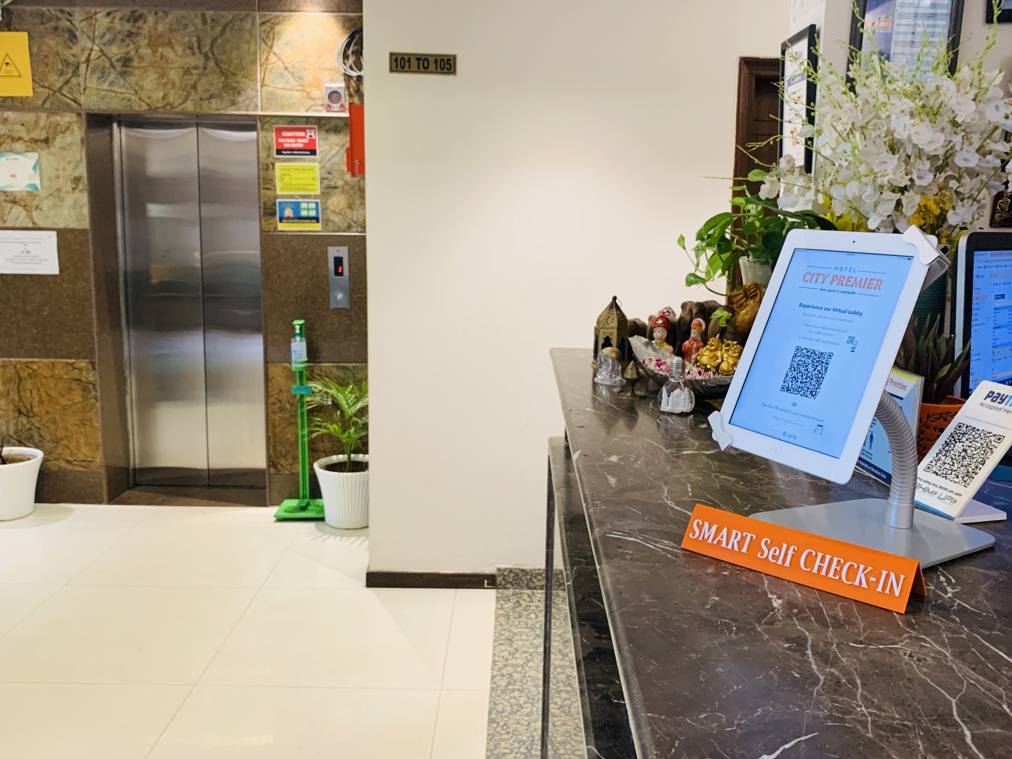 A hotel reception area with a self-check-in kiosk, a digital screen displaying hotel information, and an elevator.