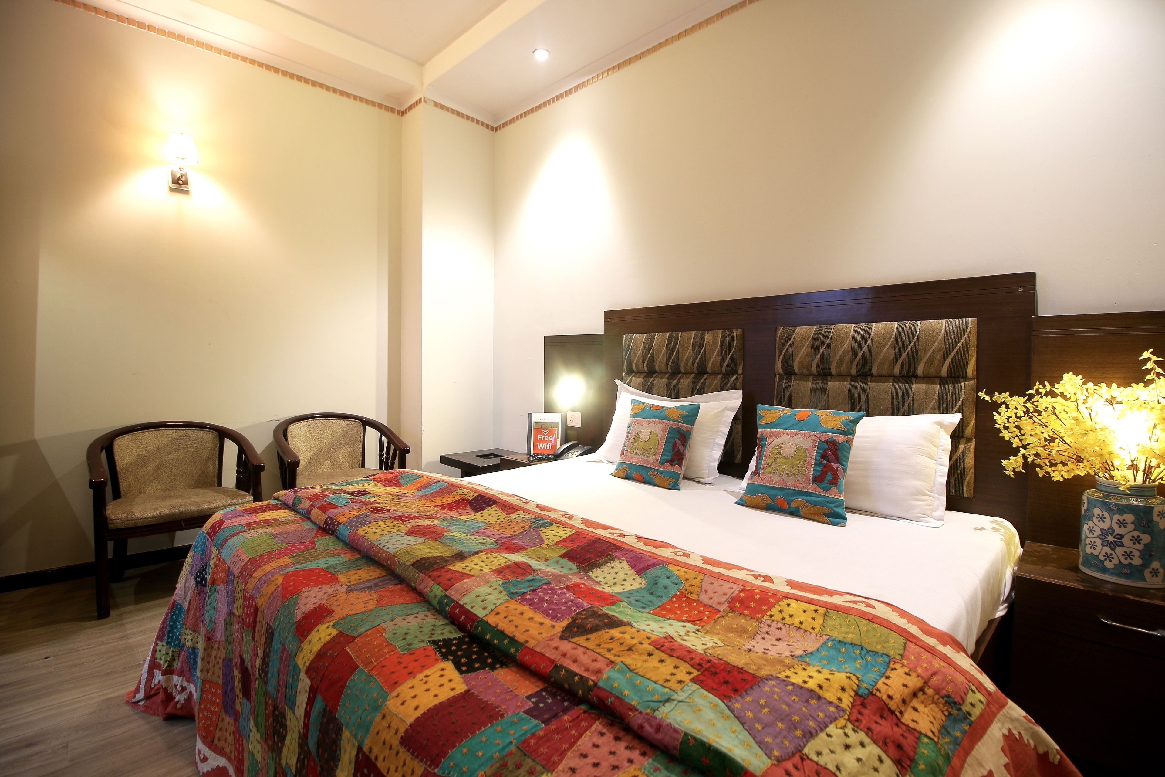 A neatly made bed with a colorful quilt, two bedside chairs, a nightstand with a lamp, and a vase with flowers on the right side.