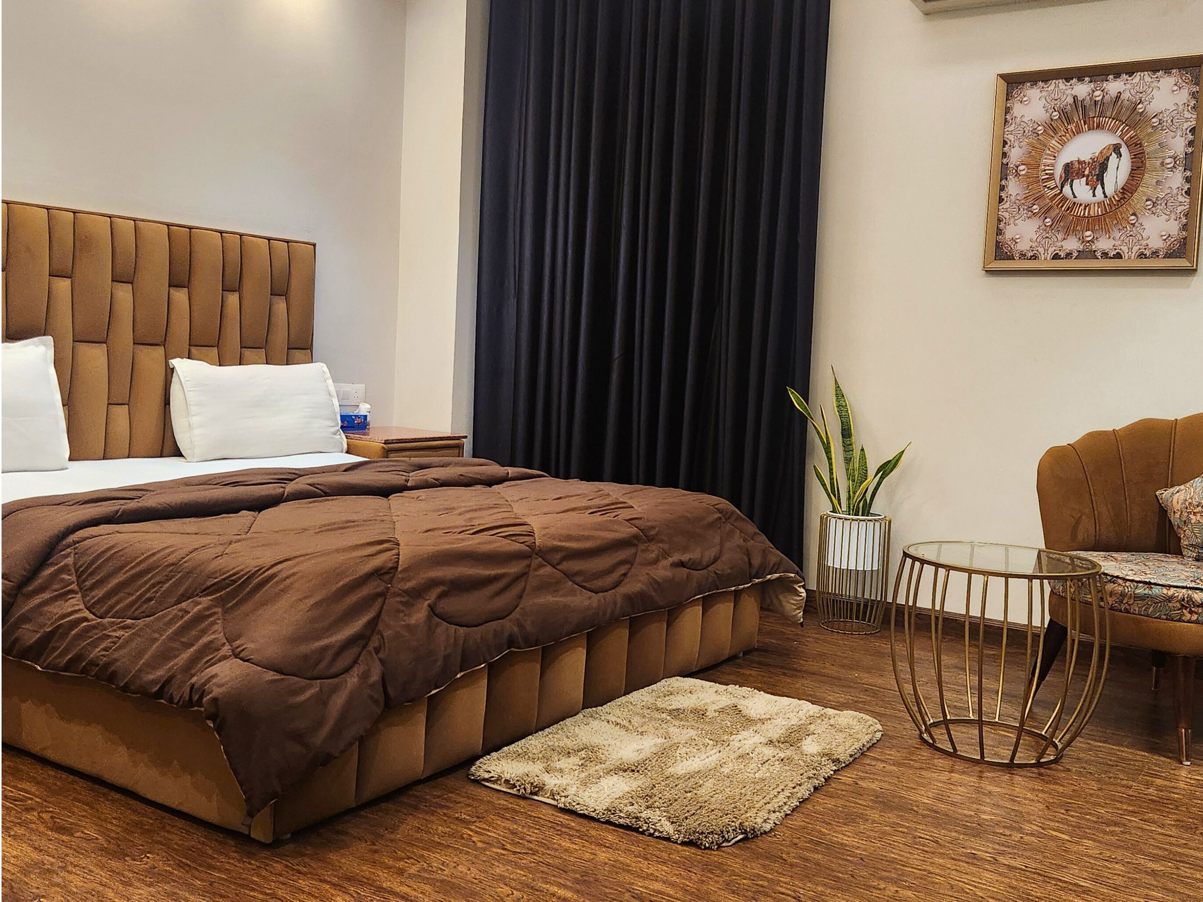 A hotel room with a large bed, a small round table, a chair, a potted plant, and a decorative wall piece.