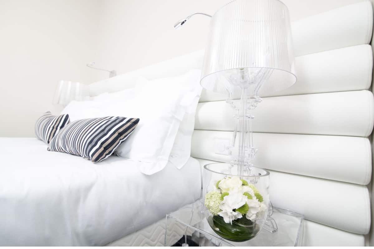Un lit blanc avec des oreillers à rayures, un vase transparent rempli de fleurs et une lampe transparente posée sur une table de chevet.