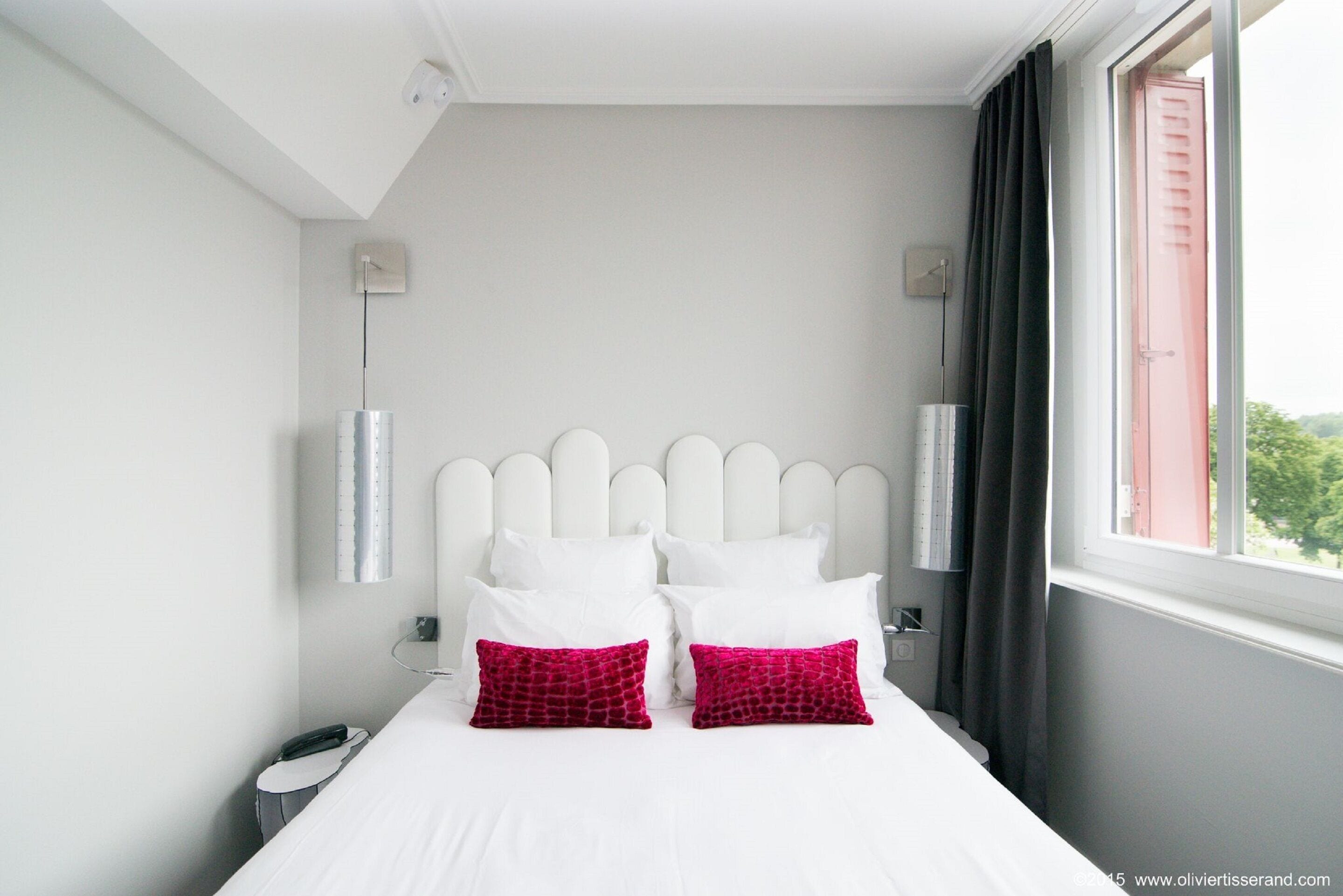 Habitación de hotel con cama blanca, dos almohadas rojas, cabecera blanca y ventana con cortinas.