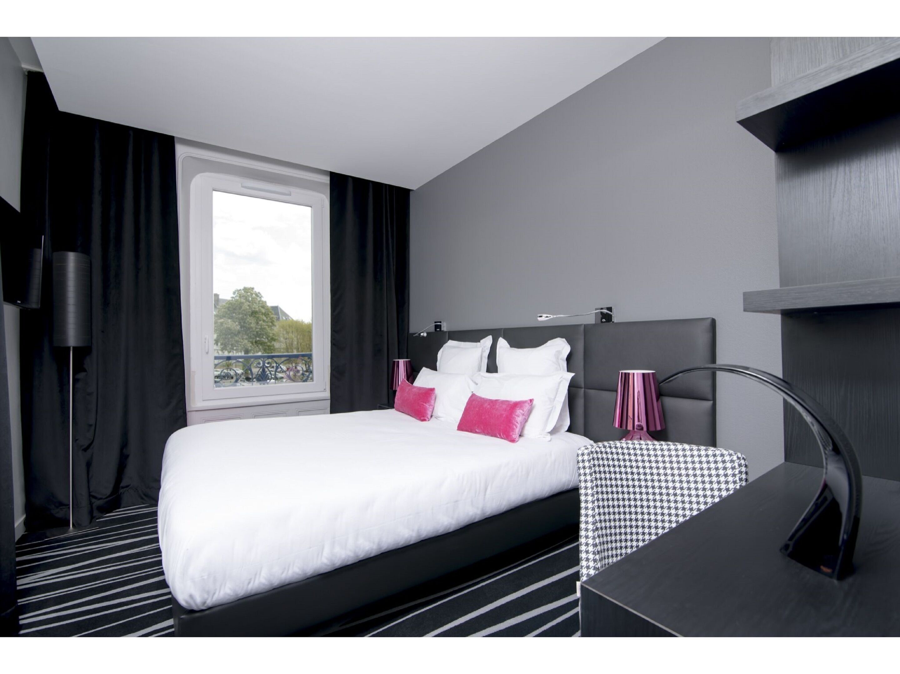 Una habitación de hotel moderna con una cama grande, alfombra a rayas negro y blanco, vistas al exterior, cabecera negra y una mesita de noche con lámpara.