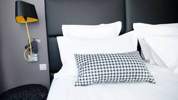 Una cama impecable con ropa blanca y una almohada de pata de gallo, una lámpara negra con pantalla amarilla y una mesita redonda con tapa estampada negra.