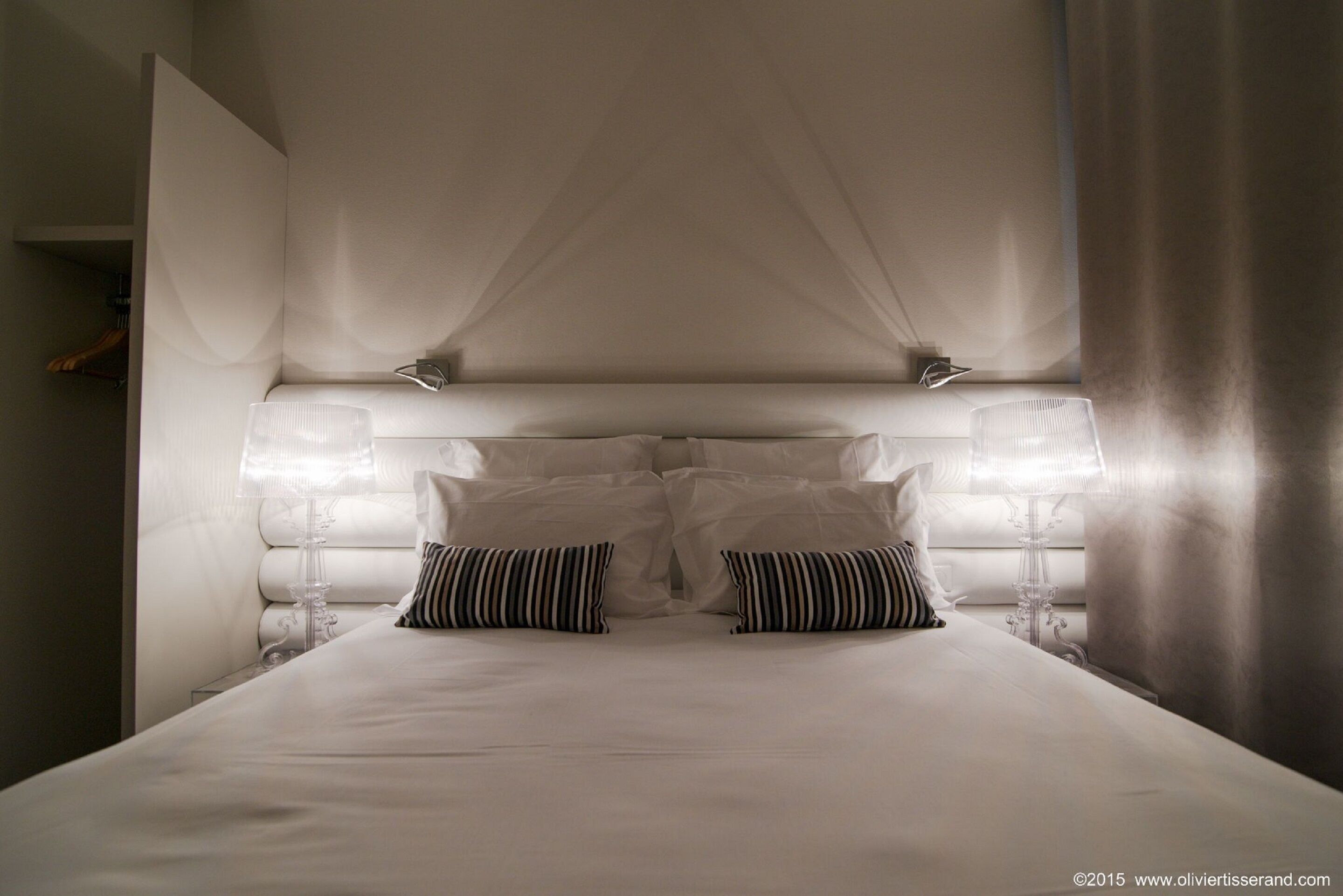 Een net opgemaakt bed met witte lakens en gestreepte kussens, een nachtkastje met een lamp, en een groot raam met gordijnen.