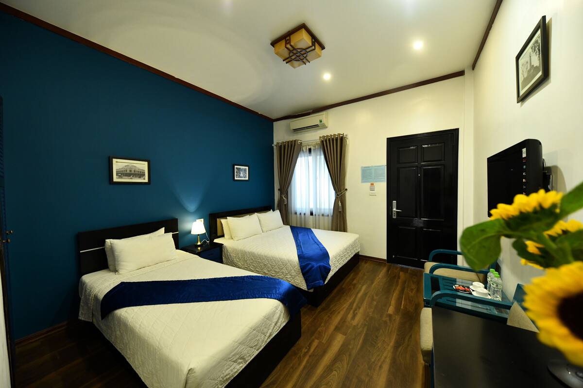 Une chambre d’hôtel avec deux lits, un bureau, une chaise, une table de chevet, un appareil de climatisation, une porte et un tableau encadré au mur.