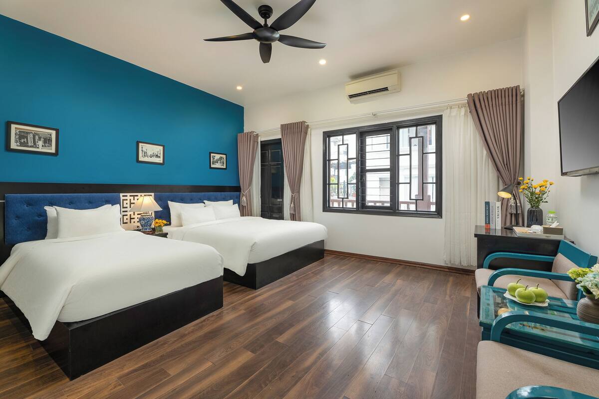 Une chambre d’hôtel avec deux lits, un ventilateur de plafond, une grande fenêtre avec des rideaux, une télévision et une petite table sur laquelle se trouvent un vase et des pommes.