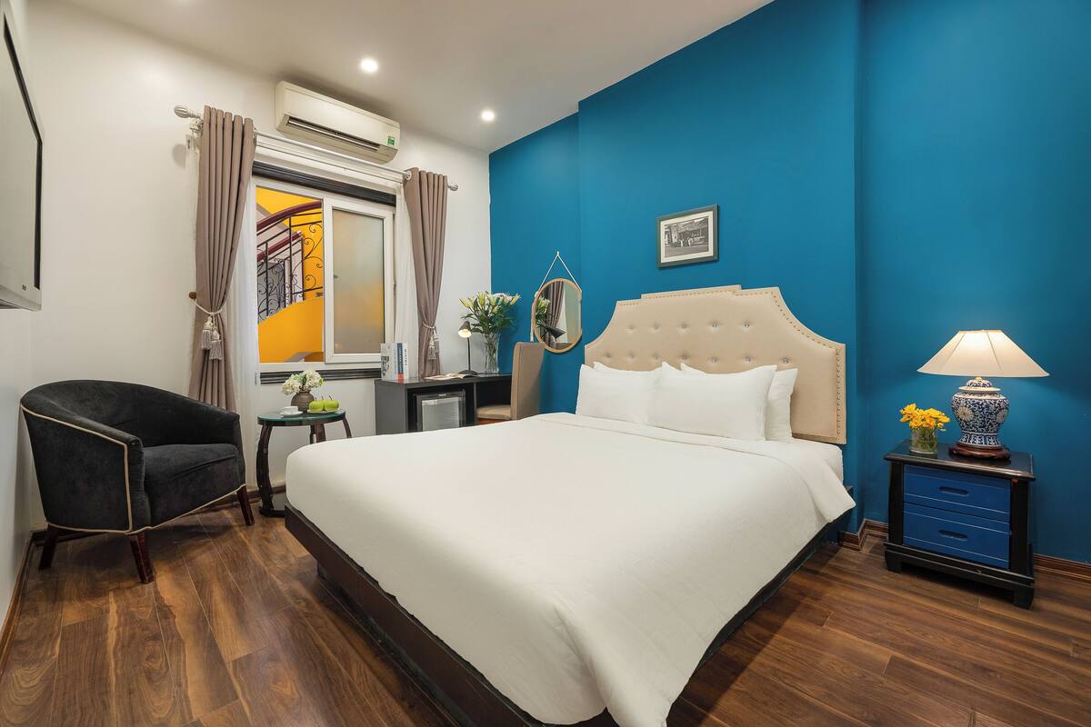 Une chambre avec un grand lit, une chaise, une table de chevet avec une lampe et une fenêtre avec des rideaux.