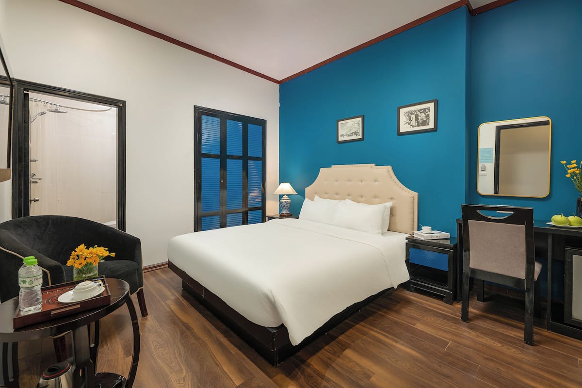Une chambre d’hôtel avec un grand lit, un bureau avec une chaise, une petite table avec une bouteille et des fleurs, et une fenêtre avec des stores.