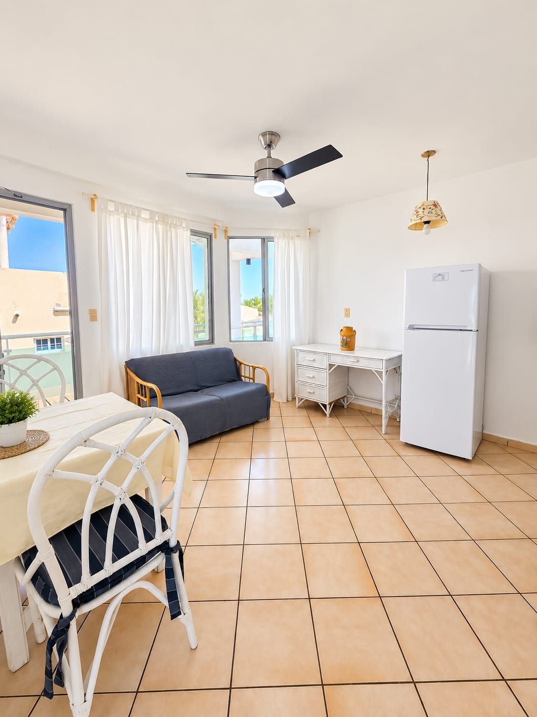 Ein kompakter Wohnbereich mit Essbereich, einer Couch, einer kleinen Küche mit Kühlschrank und einem Deckenventilator.