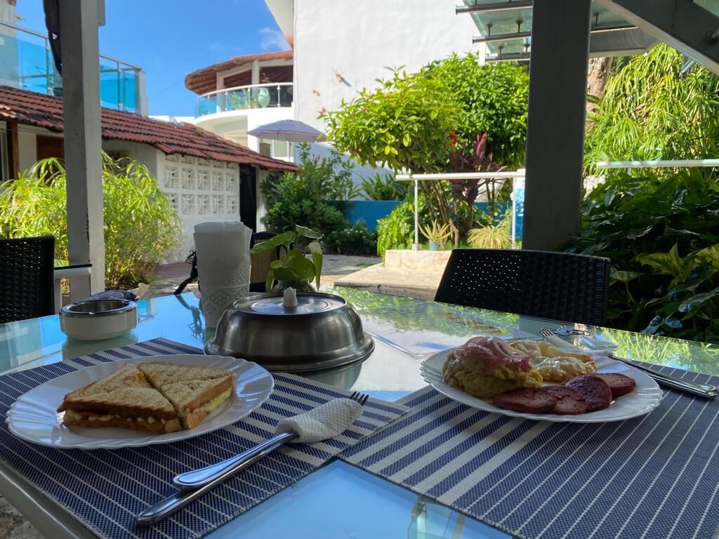 Una mesa dispuesta con alimentos para el desayuno, incluyendo sándwiches y huevos, con vista a un edificio y follaje al fondo.