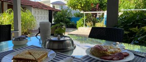 Una mesa dispuesta con alimentos para el desayuno, incluyendo sándwiches y huevos, con vista a un edificio y follaje al fondo.