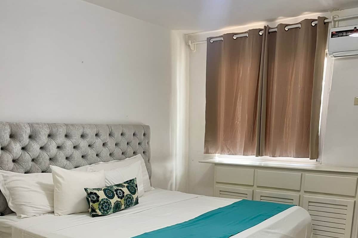Ein Schlafzimmer mit einem großen Bett, einem grauen, gepolsterten Kopfteil, weißer Bettwäsche, einer türkisblauen Decke und einem Fenster mit Vorhängen.