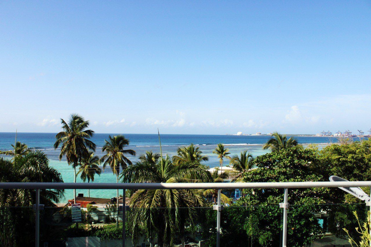 Un balcone con vista sull'oceano, palme e strutture industriali in lontananza.