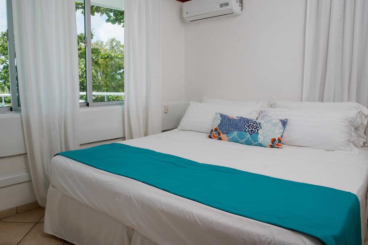 Ein ordentlich bezogenes Bett mit einem türkisblauen Überwurf und dekorativen Kissen, ein Fenster mit weißen Vorhängen und eine Klimaanlage an der Wand.