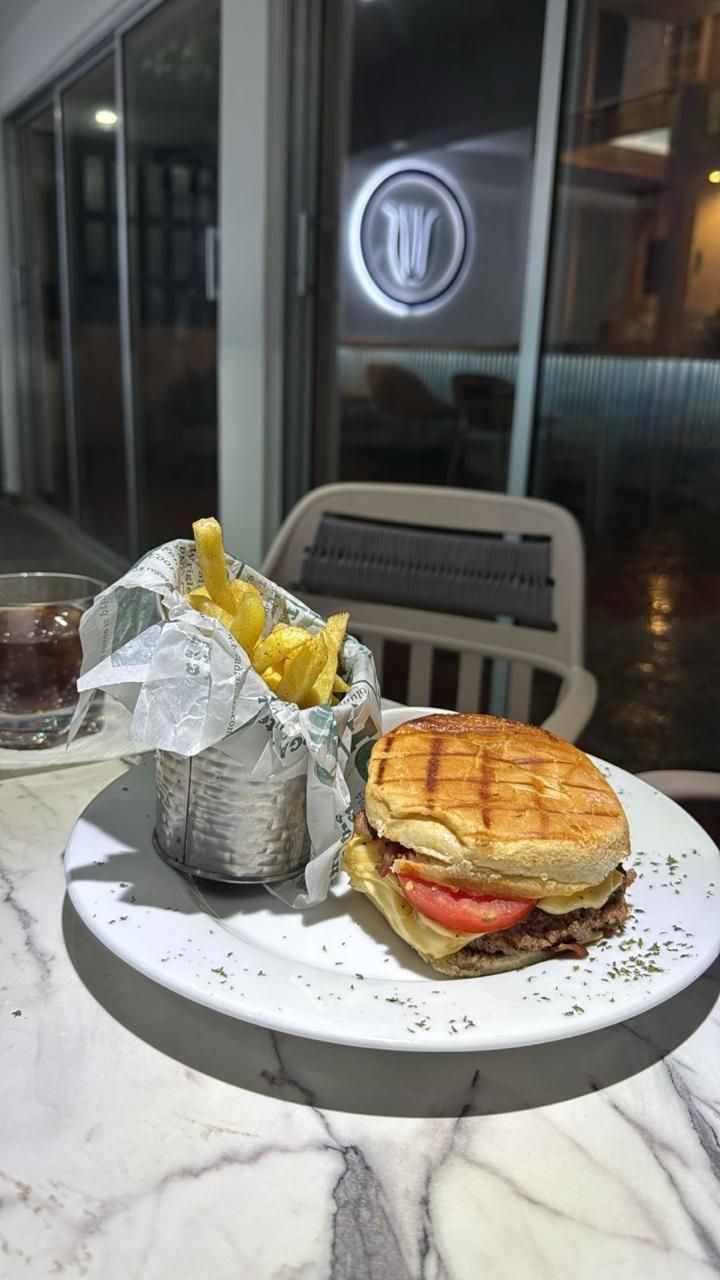 Un plato con un sándwich a la plancha y papas fritas sobre una mesa de mármol, una copa de una bebida oscura y una silla blanca al fondo.
