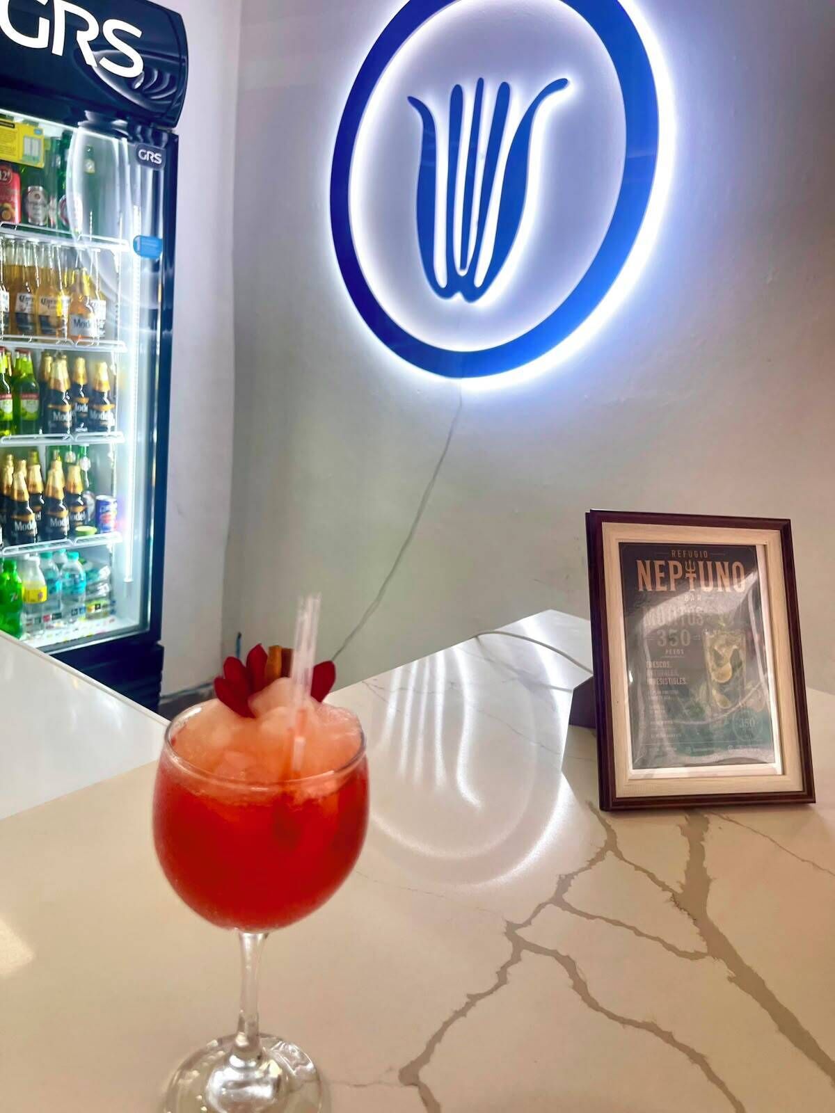 A bar area with a neon sign, a refrigerator stocked with beverages, and a framed picture on the counter.