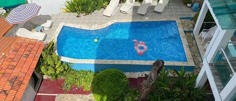 Una piscina en la azotea con agua azul cristalina, rodeada de sillones de descanso y abundante vegetación.