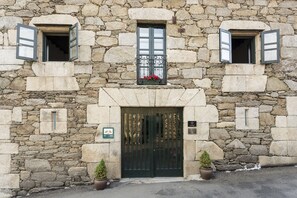 Un edificio de piedra con una entrada central, dos ventanas con contraventanas y una puerta con reja de metal.
