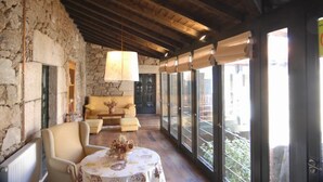 A cozy room with a stone wall, wooden ceiling beams, a sofa, a table with a floral tablecloth, and large windows with a view of the outside. - Casa Grande de Cristosende (A Teixeira)