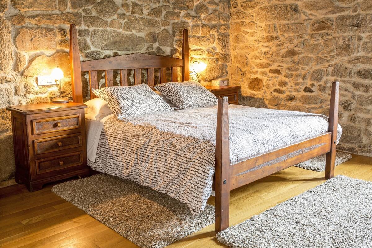 A wooden four-poster bed with a knitted blanket, two bedside tables with lamps, and a stone wall background.