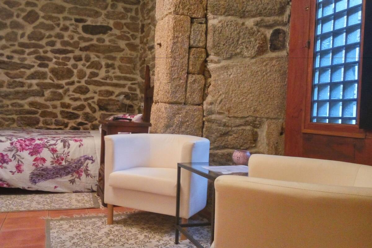 A room with a stone wall, a bed with floral bedding, a white armchair, and a window with bars.