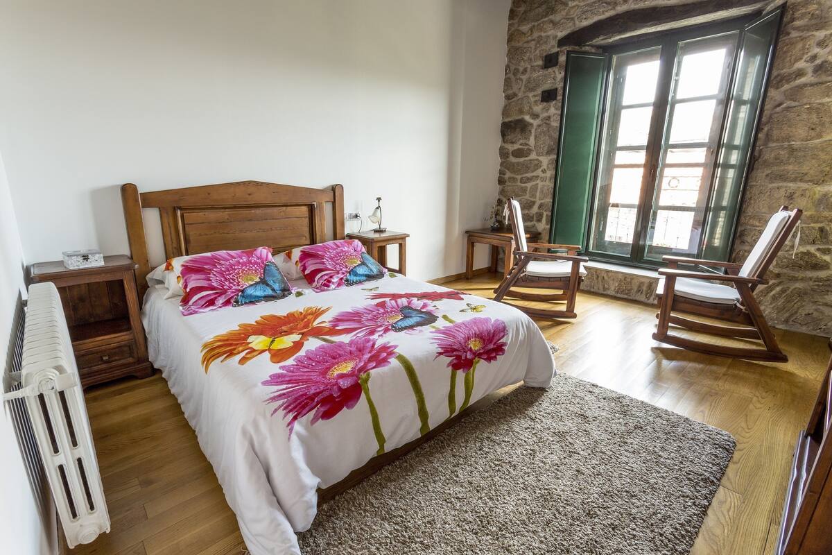 A bedroom with a wooden bed, floral bedding, a stone wall, and a rocking chair.