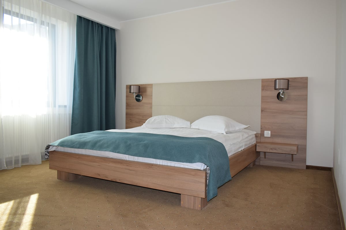 A neatly made bed with a wooden frame and a teal blanket, a bedside table with a lamp, and a window with sheer curtains.