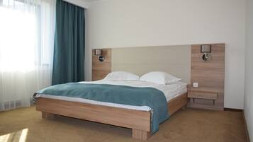 Un letto ben rifatto con struttura in legno e coperta color petrolio, un comodino con una lampada e una finestra con tende trasparenti.