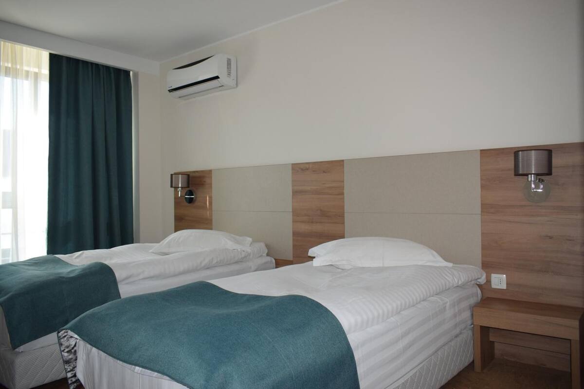 A hotel room with two beds, a window with curtains, an air conditioning unit, and wall-mounted lamps.