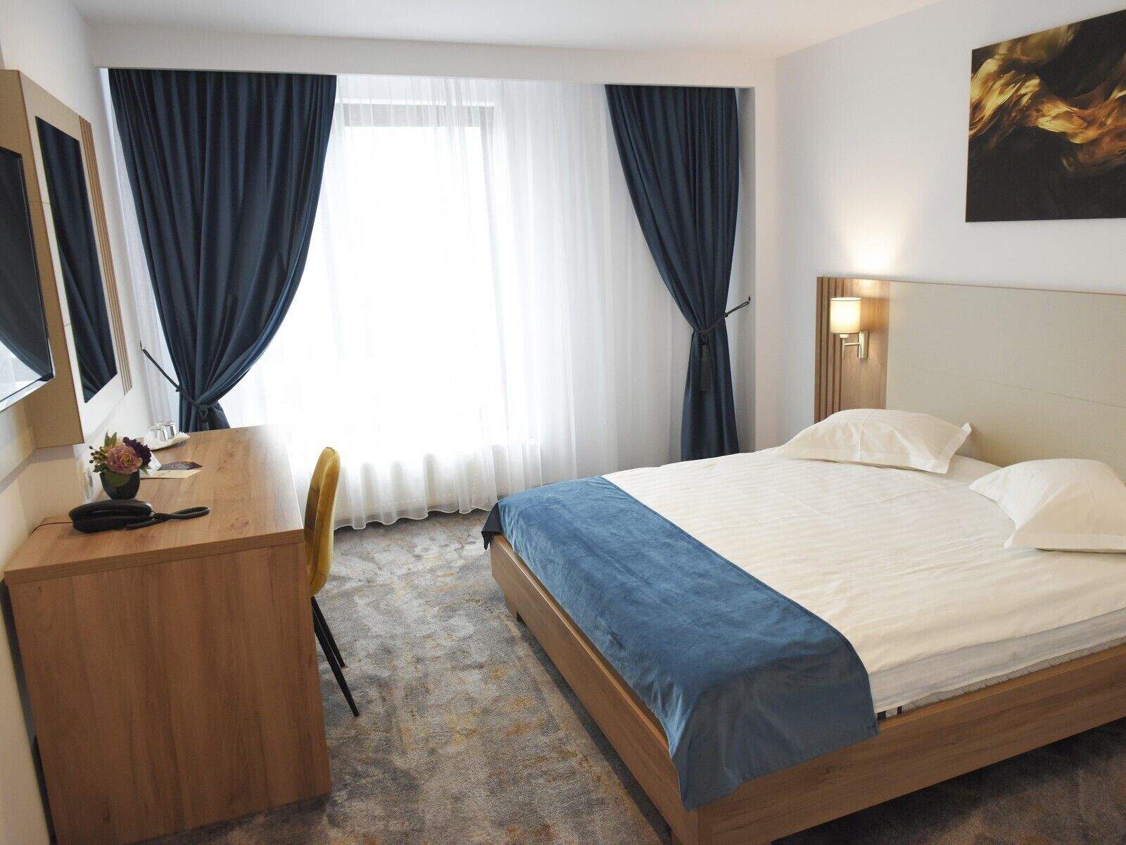 A hotel room with a bed, a desk, a chair, a telephone, a lamp, and a painting on the wall.