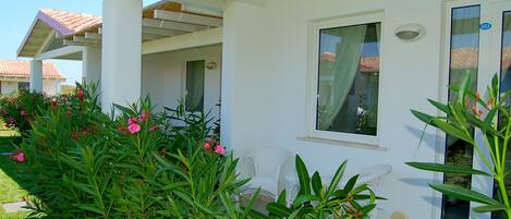 A white building with a covered patio area, a white chair, and a window with a curtain.