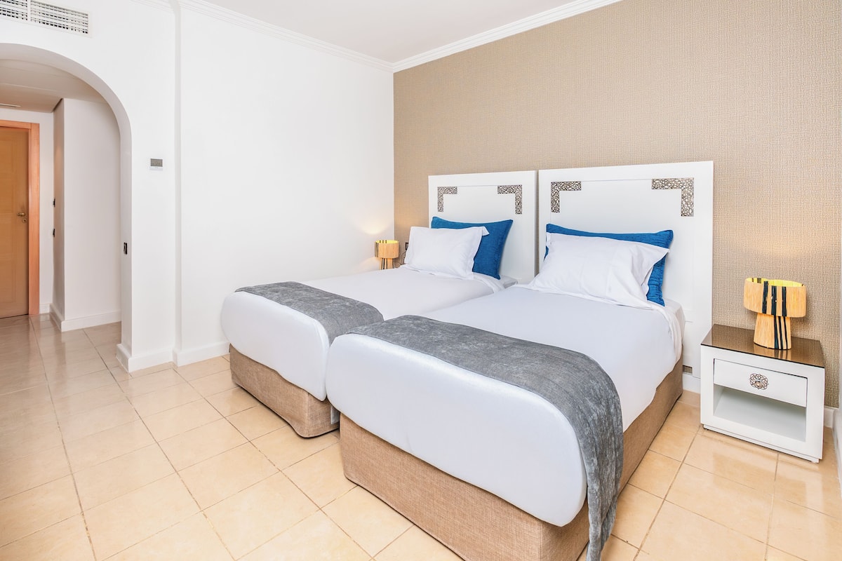 Habitació d'hotel amb dos llits, una tauleta de nit amb una làmpada i una porta de fusta.