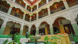 A courtyard with ornate arches and a fountain, surrounded by lush greenery and a table set for a meal.