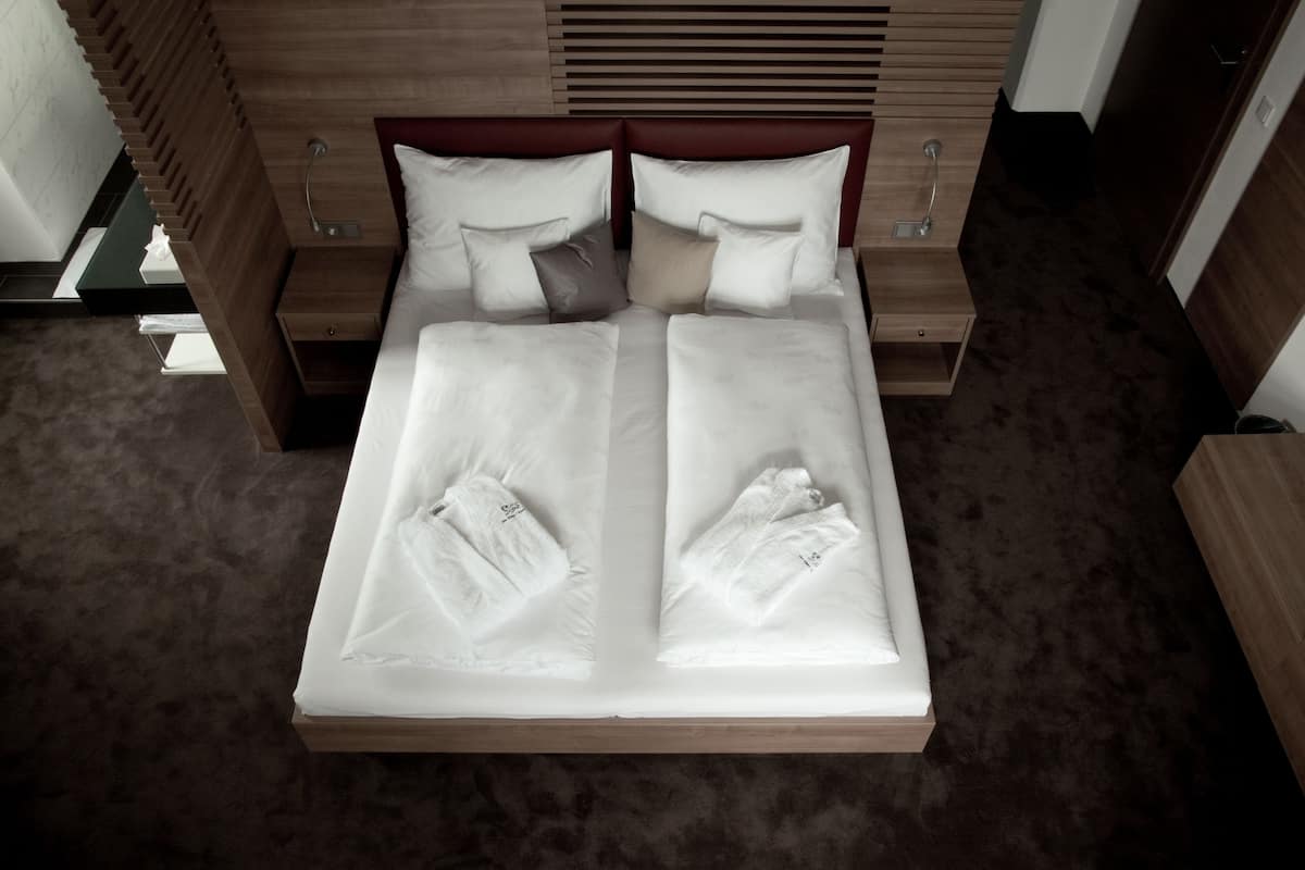 Una cama doble con sábanas blancas y dos almohadas, un cabecero de madera y una mesita de noche con una lámpara.