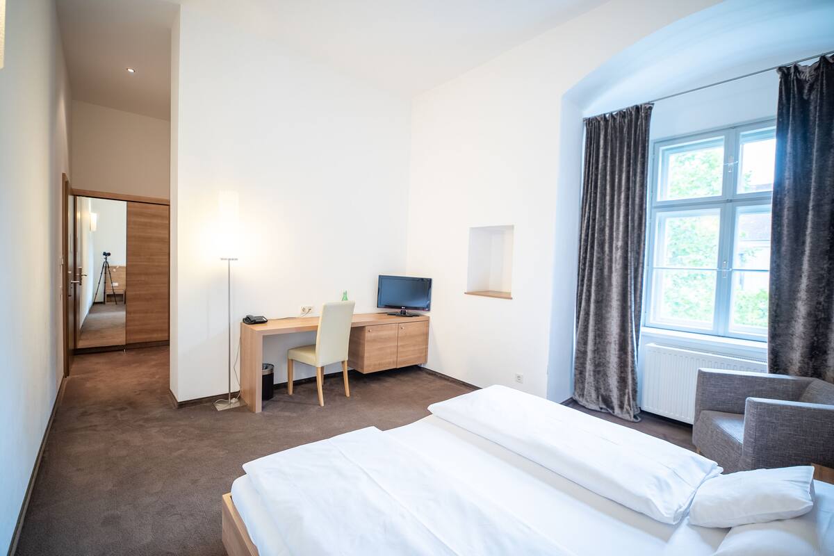 Una habitación de hotel con una cama, un escritorio con computadora, una silla y un ventanal con cortinas.