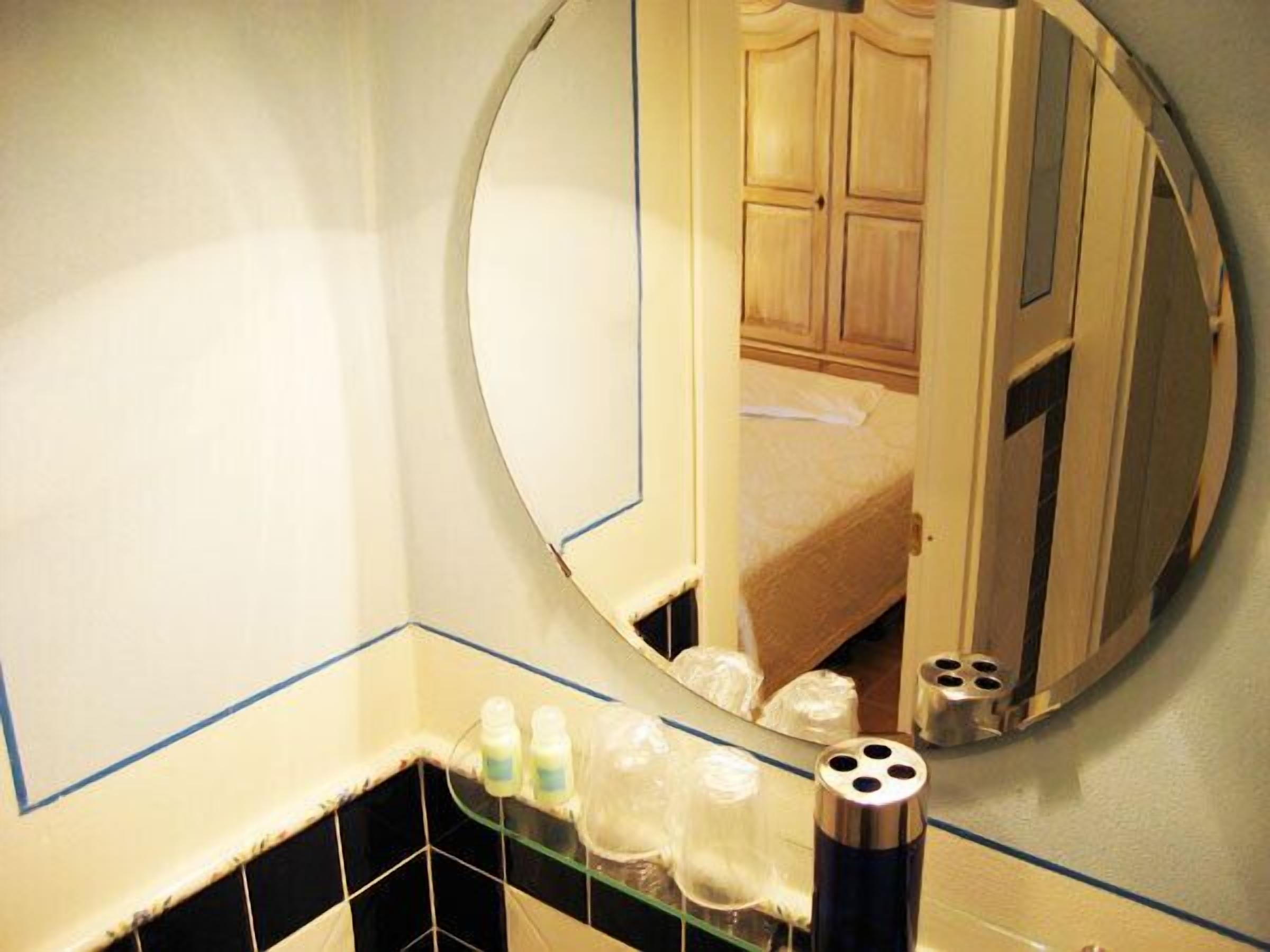 A bathroom with a round mirror, a sink with toiletries, and a visible bed in the reflection.