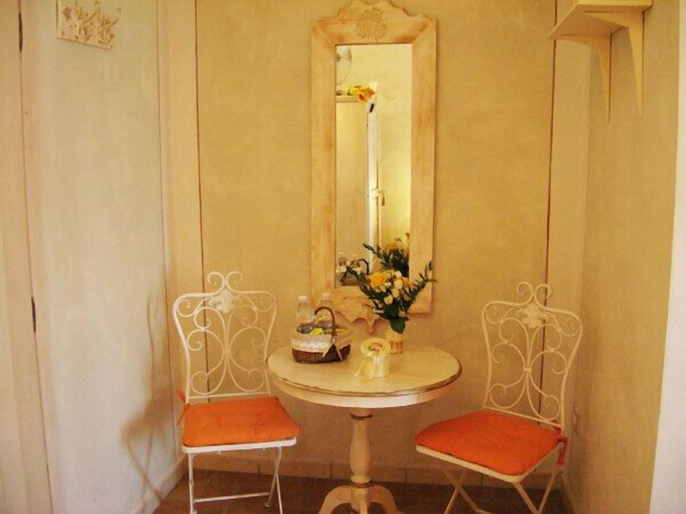 A small room with a round table, two chairs with orange cushions, a mirror, and a vase with flowers.