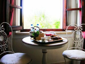 Une table avec un plateau de pâtisseries, une théière, des tasses et des bouteilles d’eau, placée près d’une fenêtre donnant sur un paysage verdoyant.